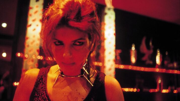 Frau mit hochgestecktem Haar und schwarzem ärmellosem Oberteil vor einer Wand mit Lichterketten und Dekorationen in warmem Licht