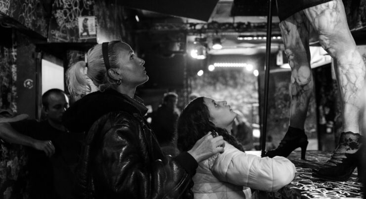 Zwei Personen blicken in einem dunklen Raum auf eine Person in High Heels auf einer erhöhten Fläche