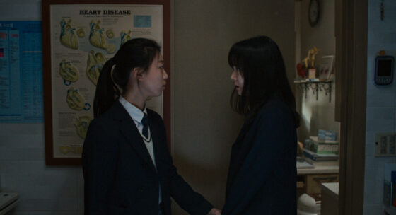 Zwei dunkel gekleidete Personen stehen sich in einem schwach beleuchteten Raum gegenüber, im Hintergrund ein Poster mit Herzdiagrammen.