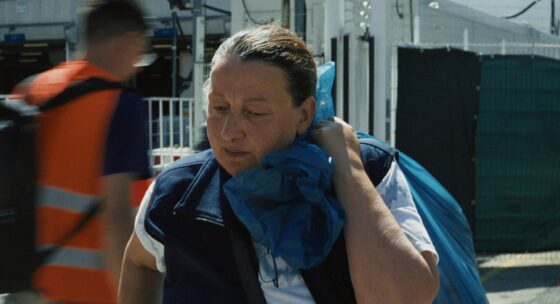 Person mit blauem Rucksack und blauem Tuch am Hals trägt etwas auf der Schulter, im Hintergrund unscharfe Person mit orangefarbener Warnweste