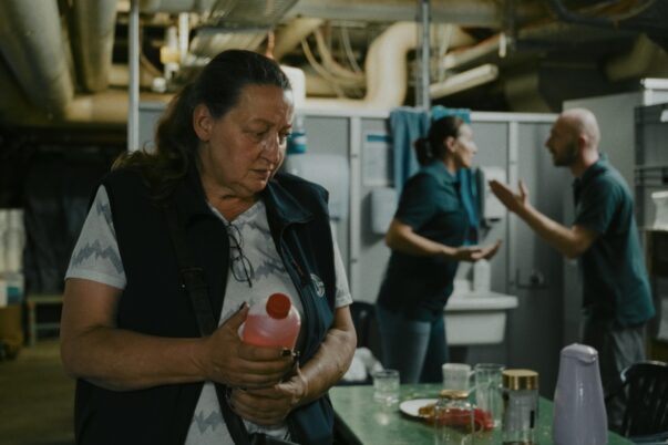 Frau hält Flasche in einem Raum mit Tisch und Geschirr, im Hintergrund streiten sich zwei Personen.