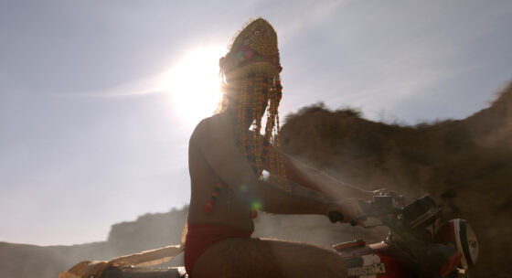 Person mit Kopfschmuck sitzt auf einem Motorrad vor der Sonne und einem Hügel