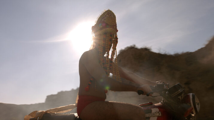 Person mit Kopfschmuck sitzt auf einem Motorrad vor der Sonne und einem Hügel