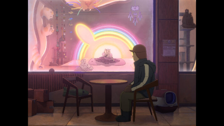 Person mit Brille und Mütze sitzt an einem runden Tisch vor einem Schaufenster mit einer leuchtenden Regenbogen-Dekoration und ausgestellten Stofftieren