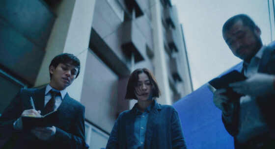 Drei Personen in Anzügen stehen vor einem Gebäude, zwei schreiben in Notizbücher, eine hält ein Tablet