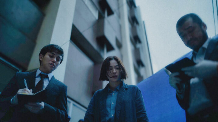 Drei Personen in Anzügen stehen vor einem Gebäude, zwei schreiben in Notizbücher, eine hält ein Tablet