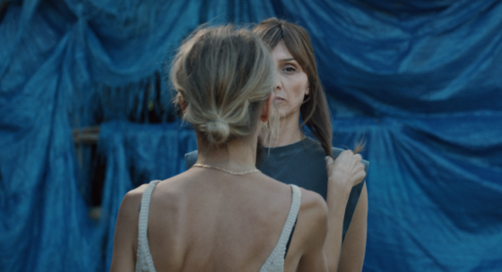 Frau mit hochgestecktem Haar und weißem gehäkeltem Top steht vor einem blauen Stoffhintergrund und hält einen Zopf einer anderen Person