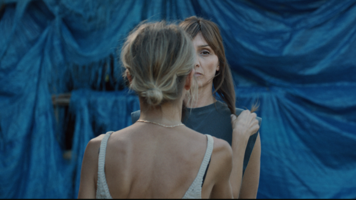 Frau mit hochgestecktem Haar und weißem gehäkeltem Top steht vor einem blauen Stoffhintergrund und hält einen Zopf einer anderen Person