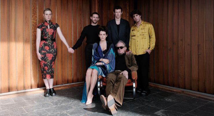 Fünf Personen vor einer Holzpaneelwand, eine Frau im gemusterten Kleid steht links, eine Frau im blauen Kleid sitzt auf einem Stuhl, drei Männer stehen, einer trägt ein gestreiftes Hemd