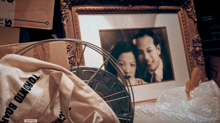 Ein vergoldeter Bilderrahmen mit einem unscharfen Porträtfoto steht in einem Keller zwischen Kartons und einer Stofftasche mit der Aufschrift 'TORONTO BOARD'.