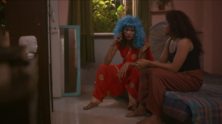Zwei Frauen sitzen in einem Zimmer, eine mit blauer Perücke und rotem Kleid auf dem Boden, die andere mit dunklen Haaren und brauner Hose auf einem Bett